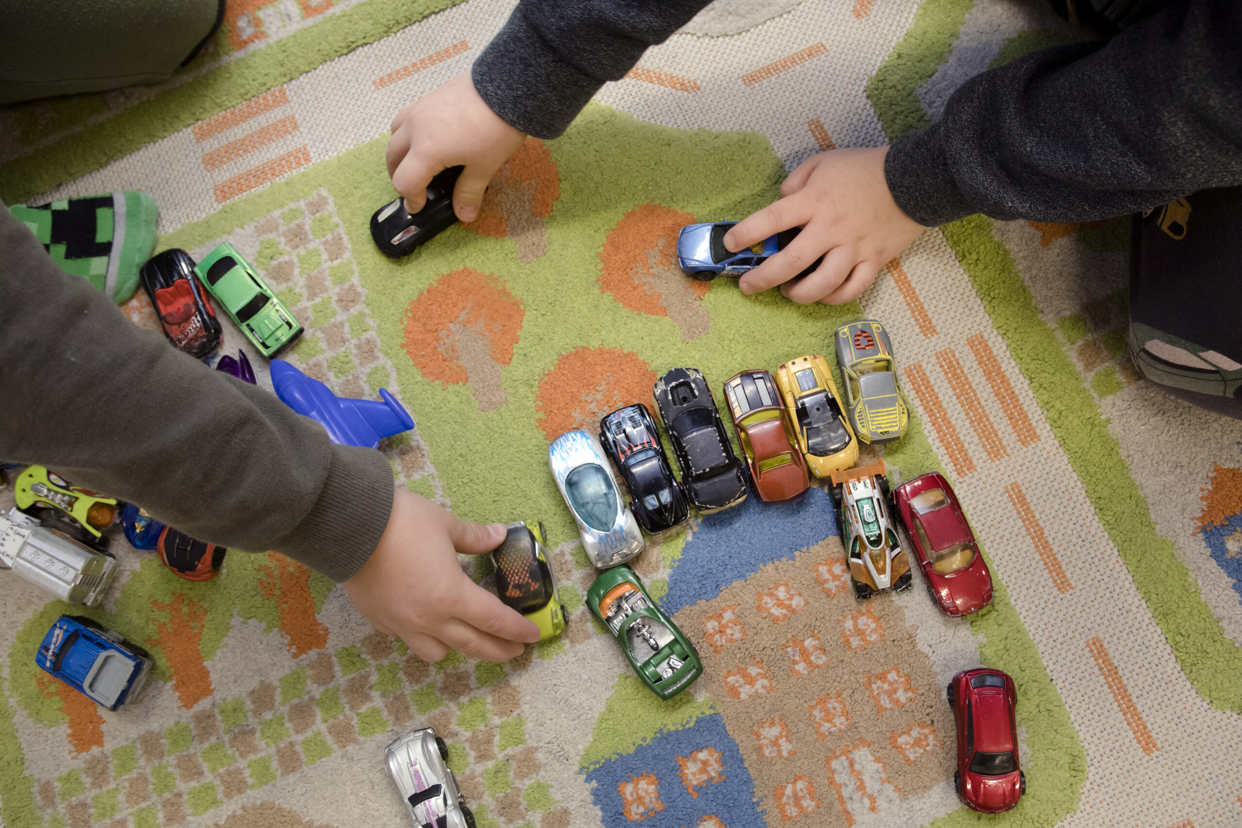 Pieniä leikkiautoja matolla, jossa ajotie- ja parkkipaikkakuvioita. Leikkivien lasten käsiä näkyvillä.