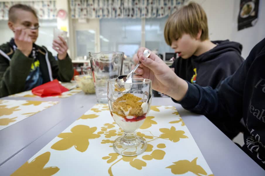 Etualalla jälkiruoka-annos ja käsi lusikoimassa sitä. Taustalla kaksi nuorta poikaa pöydän ääressä.