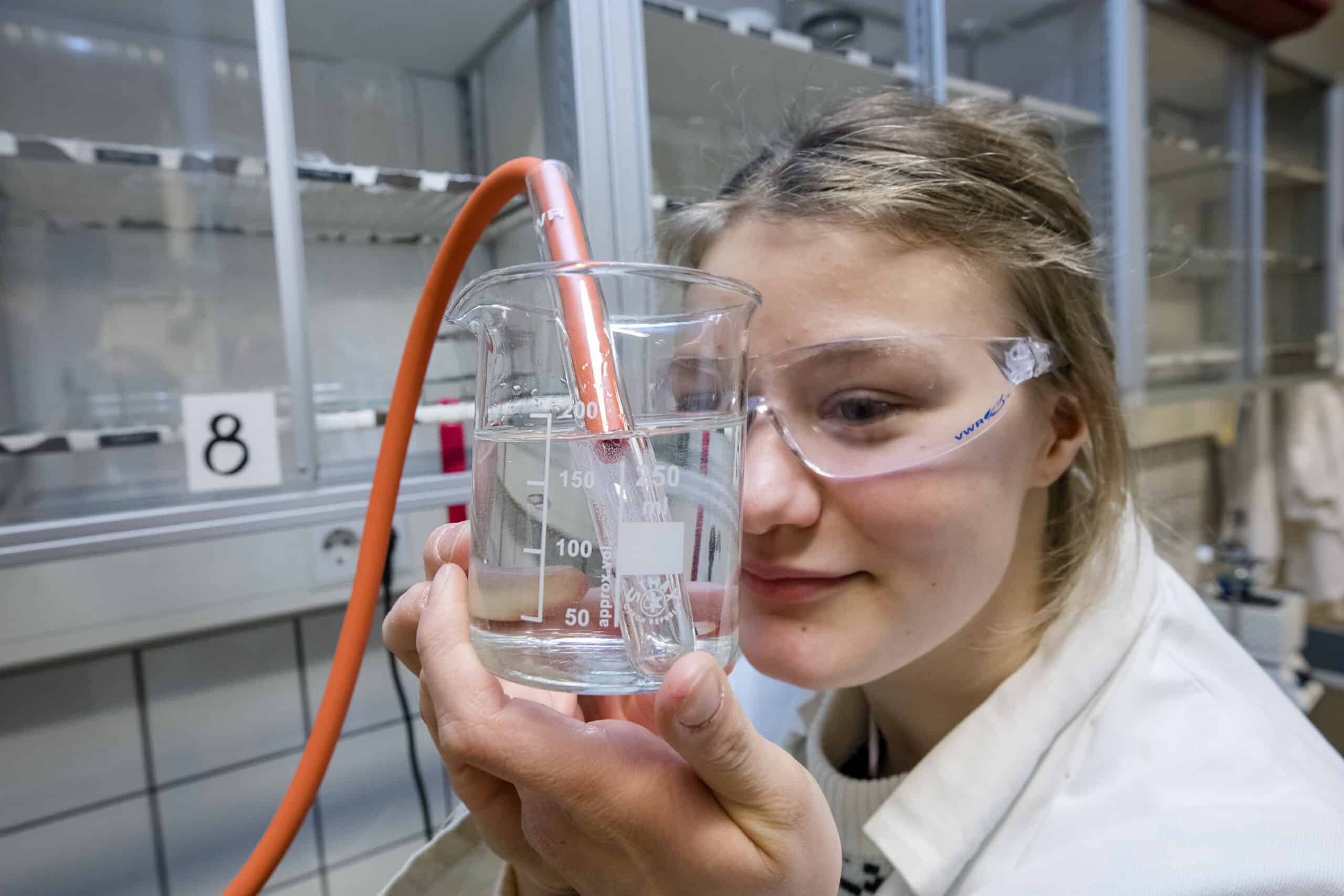 Yläkoulun oppilas kemian laboratoriossa kädessään mittalasi ja muita tarvikkeita. Suojalasit ja suojatakki. Lähikuva tytöstä.