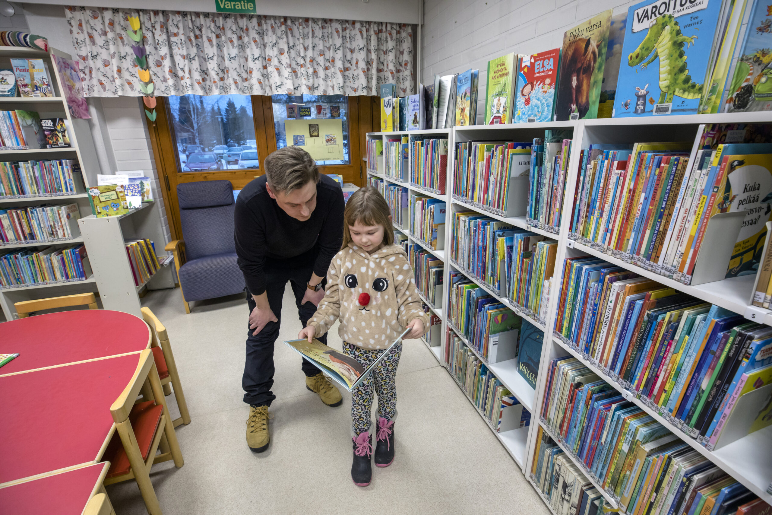 Pieni tyttö seisoo lastenosastolla kirjastossa kirja avoimena käsissään. Tytön olan takaa kirjaa katselee isä.