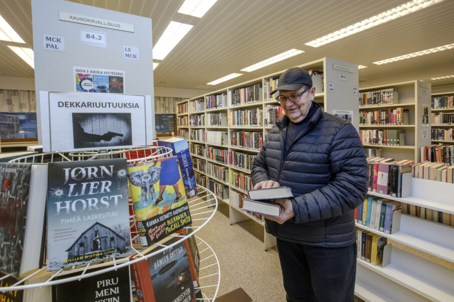 Iäkäs mies seisoo kirjastossa tutkimassa dekkarihyllyä kädessään kaksi kirjaa.