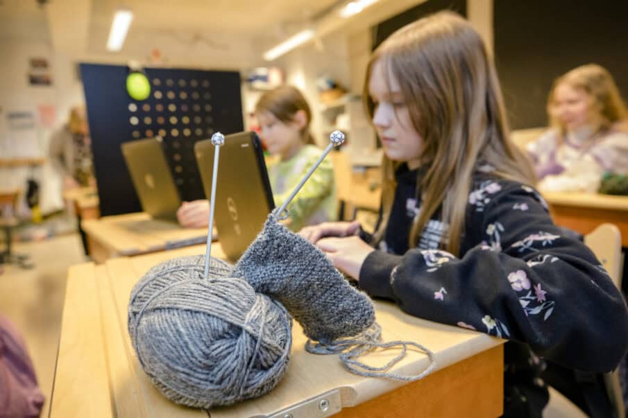 Noin yksitoistavuotias tyttö kannettavan tietokoneen äärellä koululuokassa. Etualalla lankakerä ja kudin.