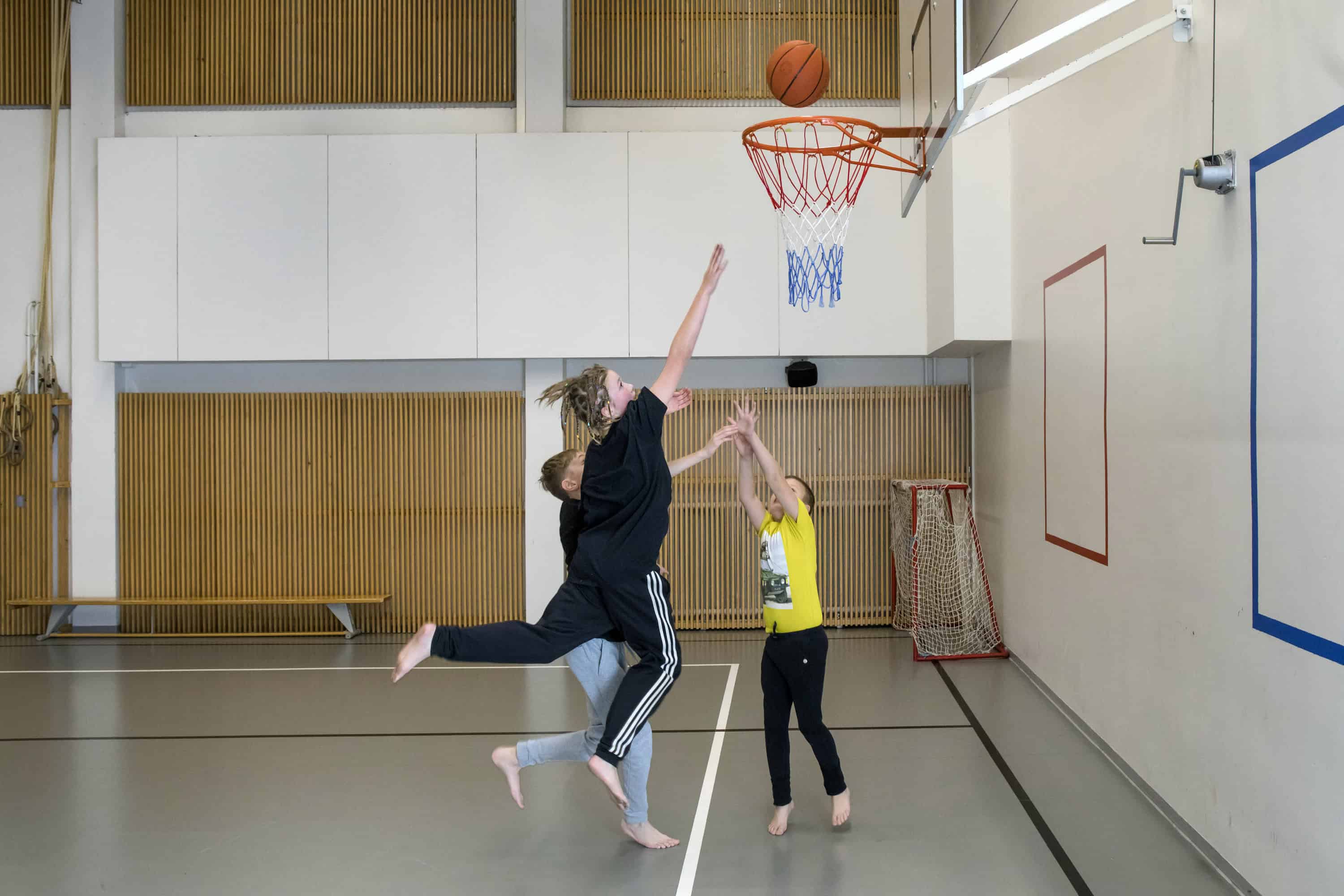 Kolme viidesluokkalaista poikaa pelaamassa koripalloa liikuntasalissa.