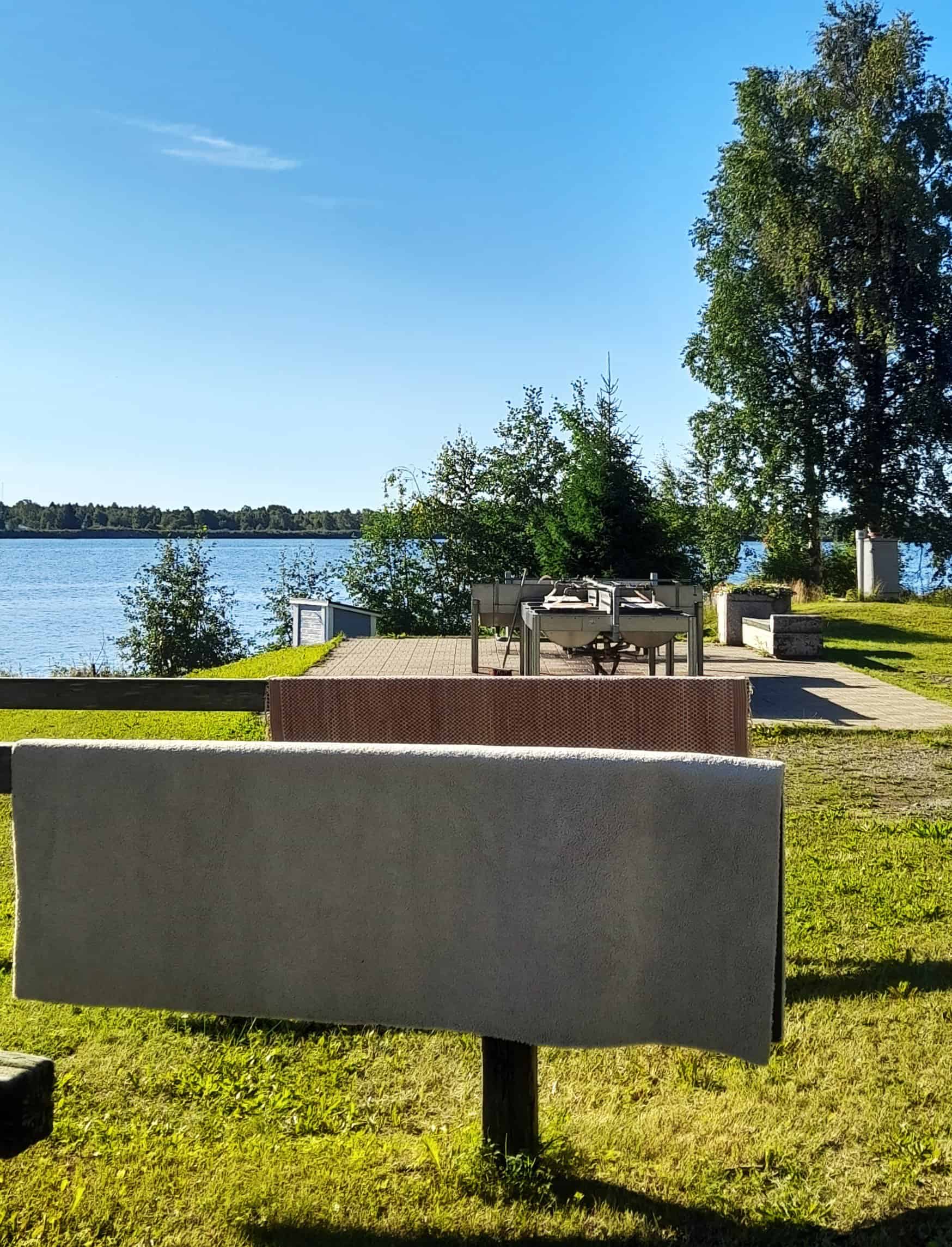 Aurinkoinen kesäpäivä, etualalla isoja mattoja kuivumassa telineiden päällä. Taustalla joki sekä terassi, jonka keskellä matonpesualtaita.