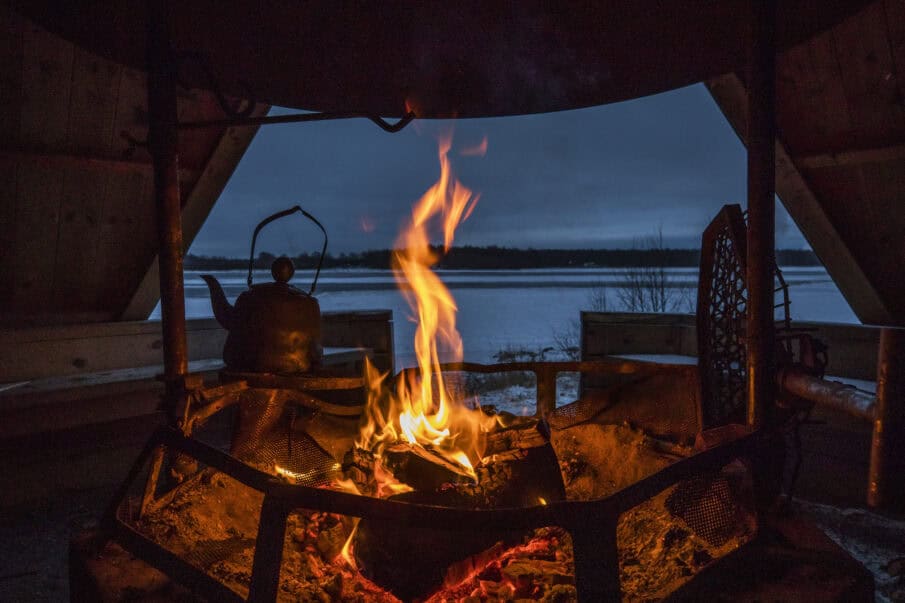 Laavun sisätiloja. Etualalla palava nuotio ja sen yllä kahvipannu. Oviaukosta näkyy lumista maisemaa.