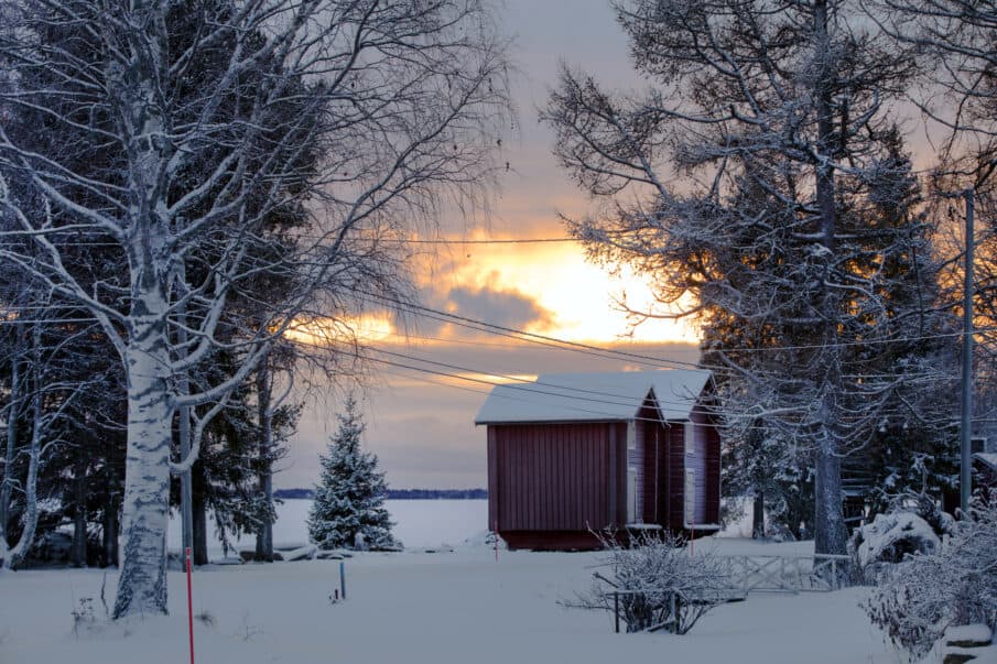 Talvinen maisema. Vasemmalla ja oikealla puita, keskellä kaksi pientä punaista aittarakennusta. Taustalla joki.