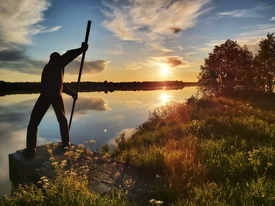 Kesäinen kuva joenrannalta. Vasemmalla lippoavaa kalastajaa esittävä patsas jalustalla. Kuvattu kohti auringonlaskua.