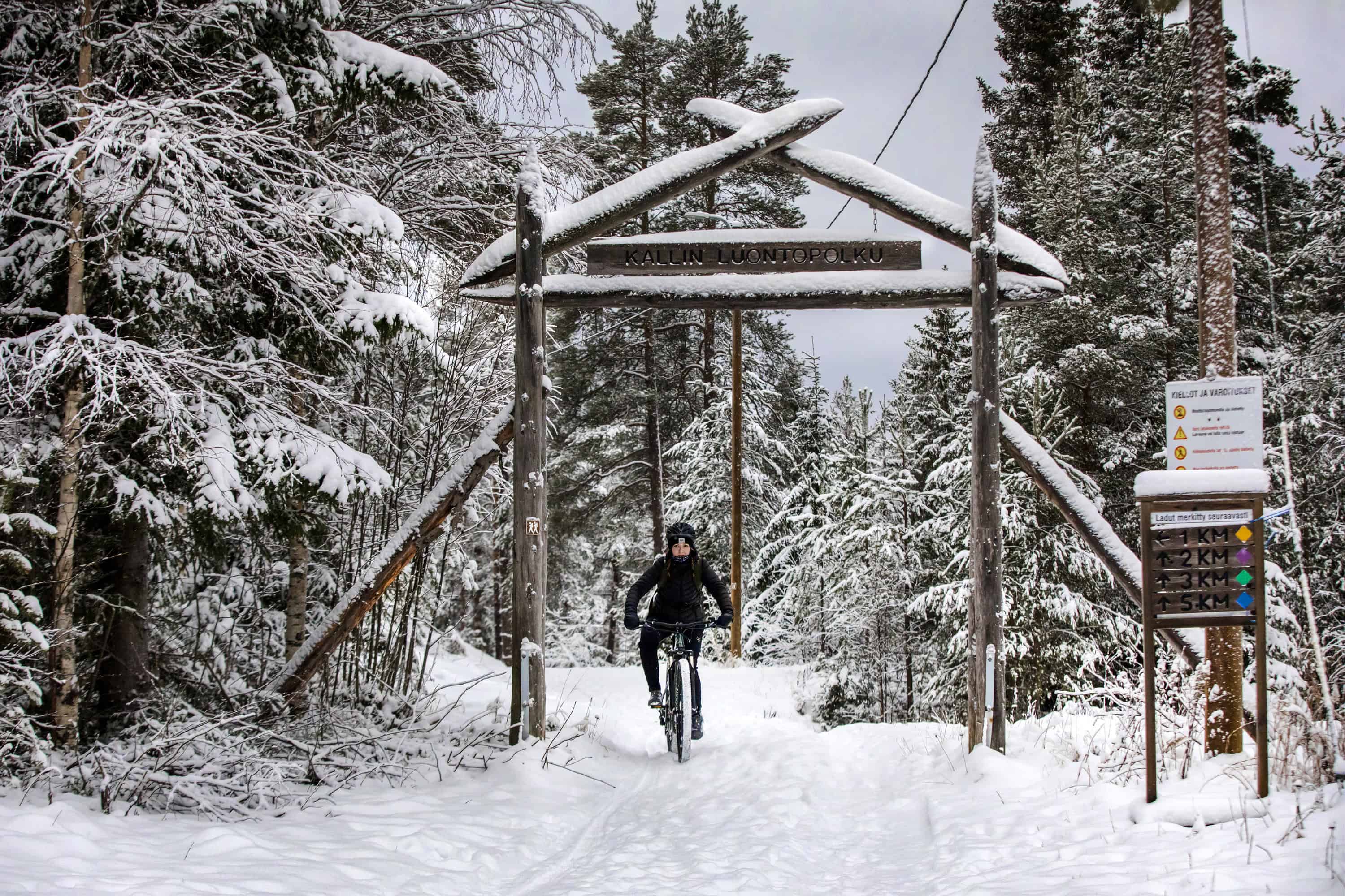 Nuori nainen ajaa maastopyörällä metsässä. Talvinen kuva. Kuvassa myös luontopolkujen reittiopasteita.