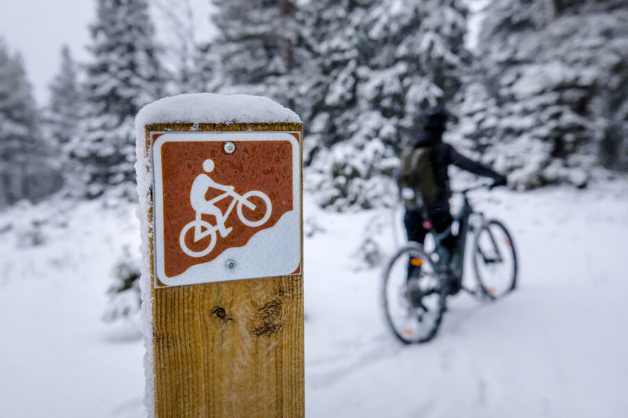 Lähikuvassa maastopyöräilyreitin merkki puupylvääseen ruuvattuna. Taustalla maastopyöräilijä taluttamassa pyöräänsä. Luminen talvinen kuva.