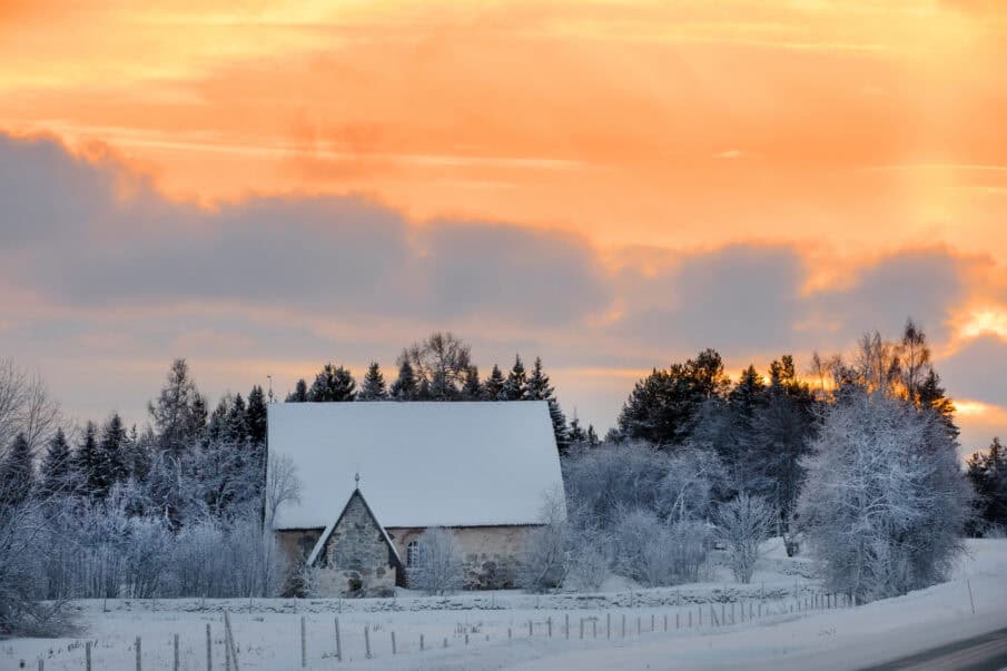 Keskiaikainen kivikirkko talvisessa maisemassa. Taivas keltainen, sinertäviä pilviä.