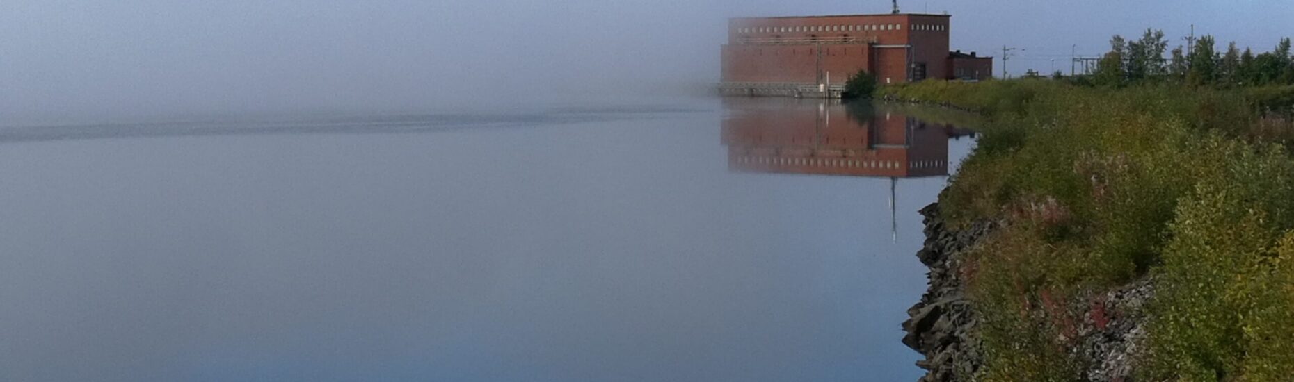 Keskellä kuvaa punatiilinen vesivoimalaitos osin sumun peittämänä. Oikealla joen rantaa.