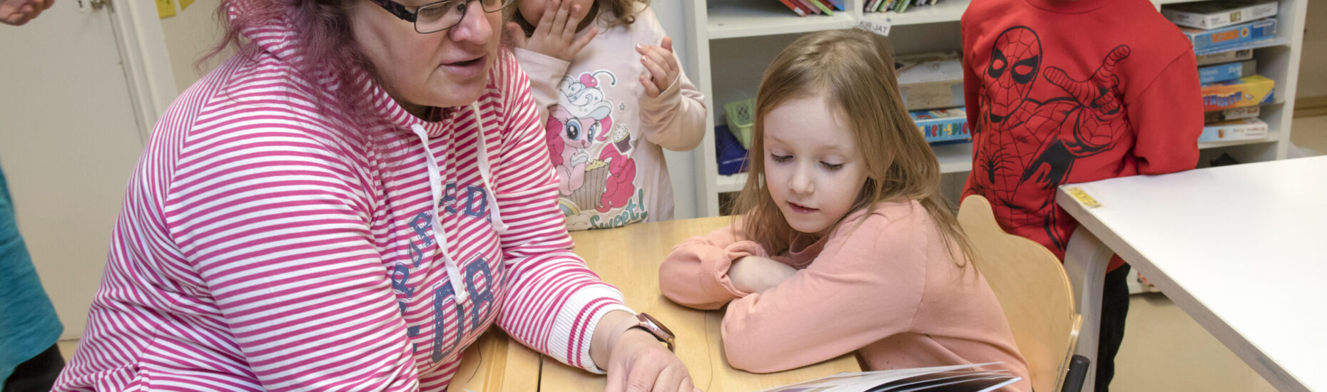 Nainen lukee kirjaa kolmelle noin 5-6-vuotiaalle lapselle luokkahuoneessa.