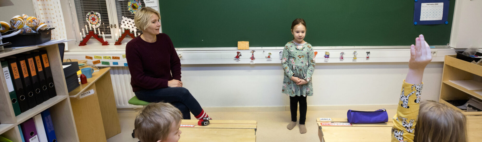 Esikoululaisten luokkahuoneessa opettaja istuu luokan edessä tuolilla. Hänen vieressään seisoo pieni tyttö ja edessään pulpeteissa istuu kaksi lasta.