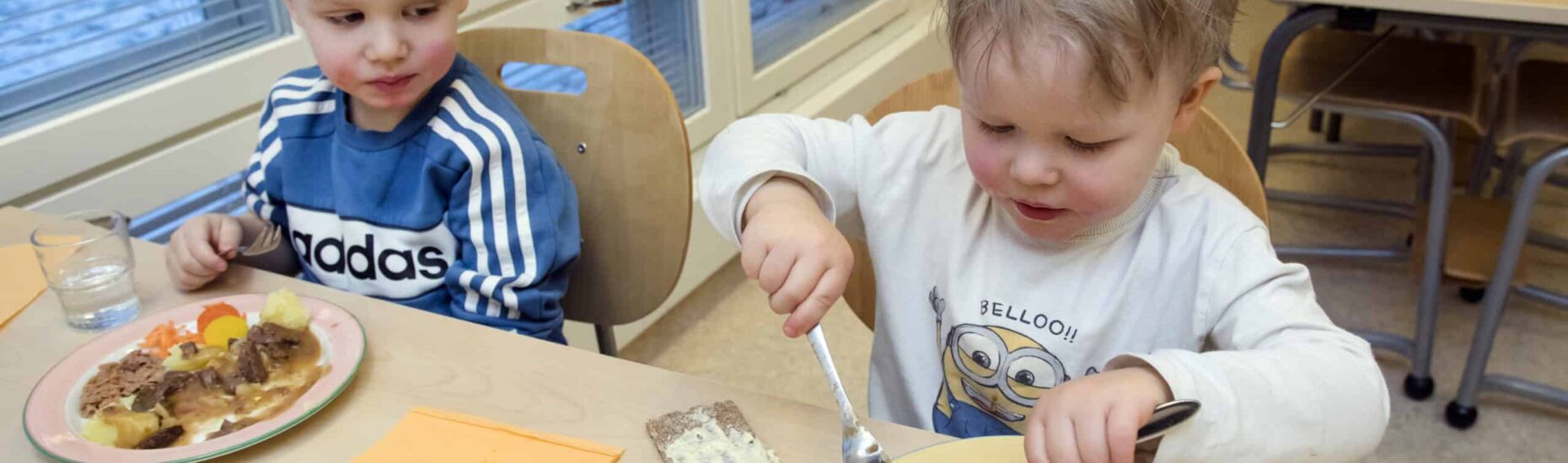 Kaksi pikkupoikaa syömässä pöydän ääressä ruokalautaset edessään.