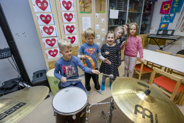 Viisi ekaluokkalaista seisoo musiikkiluokassa. Etualalla rumpuja soittava poika ja helistintä soittava poika.