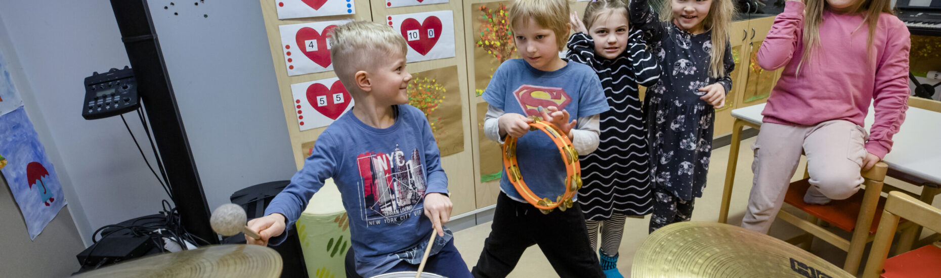 Viisi ekaluokkalaista seisoo musiikkiluokassa. Etualalla rumpuja soittava poika ja helistintä soittava poika.