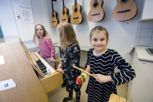 Kolme ekaluokkalaista tyttöä musiikkiluokassa. Kuvassa myös piano ja kitaroita seinällä. Etummaisella tytöllä käsissään guiro-lyömäsoitin.