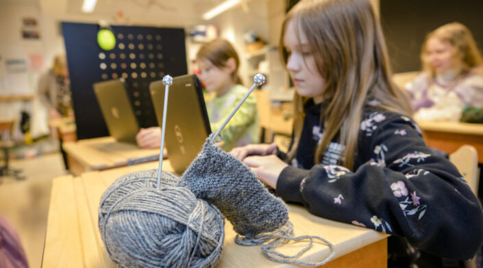 Noin yksitoistavuotias tyttö kannettavan tietokoneen äärellä koululuokassa. Etualalla lankakerä ja kudin.