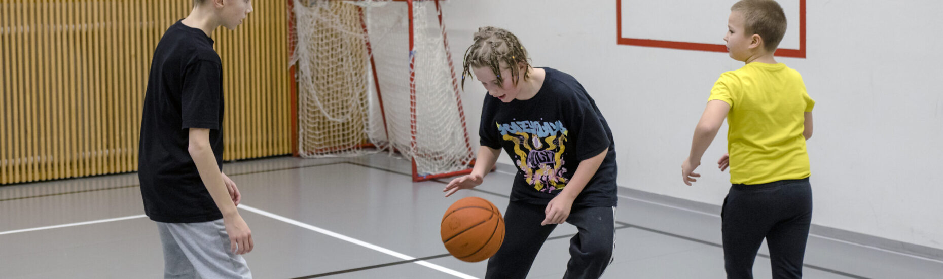 Kolme viidesluokkalaista poikaa pelaamassa koripalloa liikuntasalissa.
