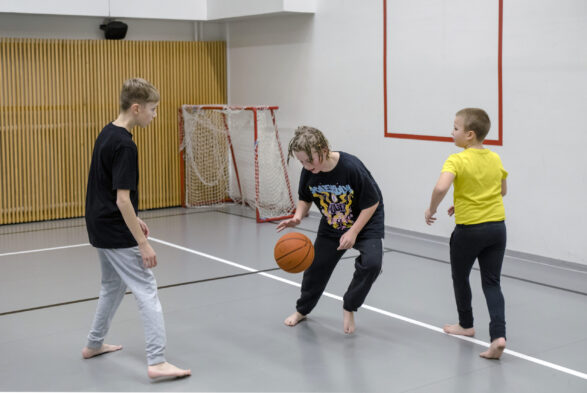 Kolme viidesluokkalaista poikaa pelaamassa koripalloa liikuntasalissa.