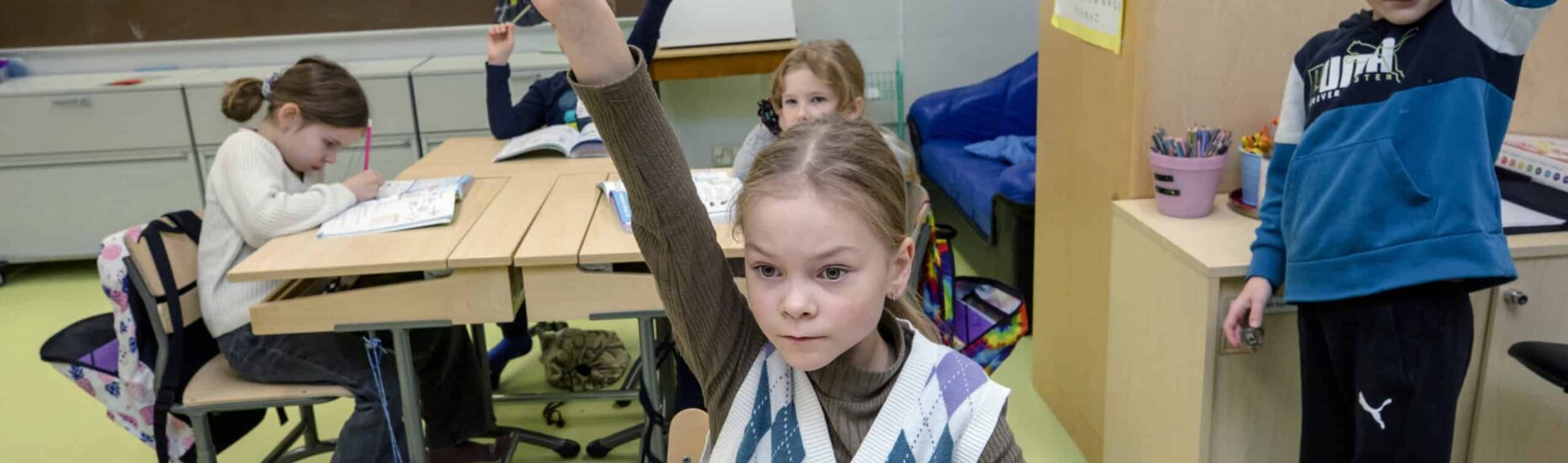 Tokaluokkalaisia luokassa. Etualalla istuva tyttö ja seisova poika, jotka viittaavat.