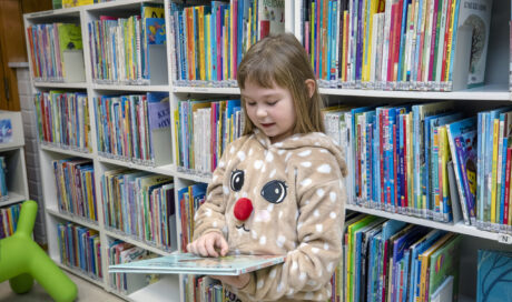 Pieni tyttö seisoo kirjaston lastenosaston hyllyn edessä kirja kädessään.