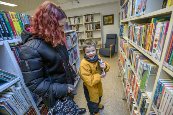 Pieni poika seisomassa äitinsä kanssa kirjaston hyllyjen välissä.