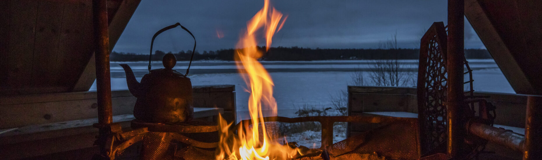 Laavun sisätiloja. Etualalla palava nuotio ja sen yllä kahvipannu. Oviaukosta näkyy lumista maisemaa.