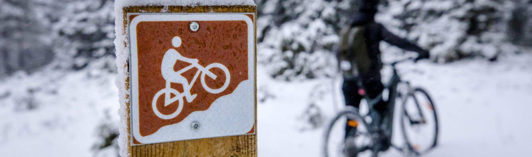 Lähikuvassa maastopyöräilyreitin merkki puupylvääseen ruuvattuna. Taustalla maastopyöräilijä taluttamassa pyöräänsä. Luminen talvinen kuva.