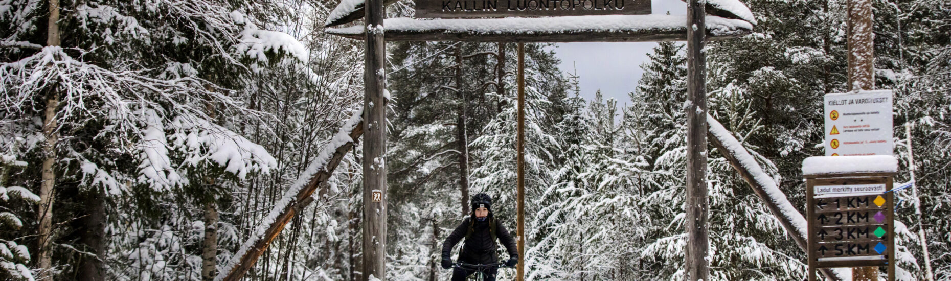 Nuori nainen ajaa maastopyörällä metsässä. Talvinen kuva. Kuvassa myös luontopolkujen reittiopasteita.