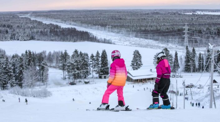 Laskettelukeskuksen rinteessä kaksi nuorta lähdössä alas. Taustalla maisemaa. Kuvattu huipulta.