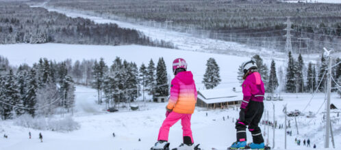 Laskettelukeskuksen rinteessä kaksi nuorta lähdössä alas. Taustalla maisemaa. Kuvattu huipulta.