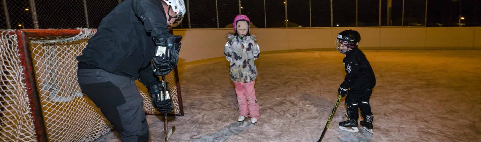 Isä pelaamassa kaukalossa jääkiekkoa pojan ja tytön kanssa.