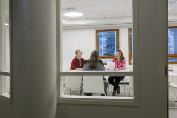 Näkymä ikkunoiden takaa kokoushuoneeseen, jossa istuu kolme naista.