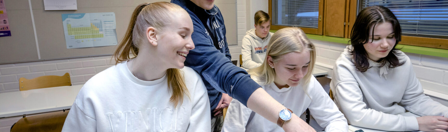 Lukiolaisia oppitunnilla. Eturivissä kannettavien tietokoneiden ja pulpettien ääressä kolme tyttöä, taustalla yksi poika. Tyttöjen takana seisoo miesopettaja.