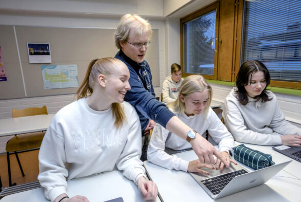 Lukiolaisia oppitunnilla. Eturivissä kannettavien tietokoneiden ja pulpettien ääressä kolme tyttöä, taustalla yksi poika. Tyttöjen takana seisoo miesopettaja.