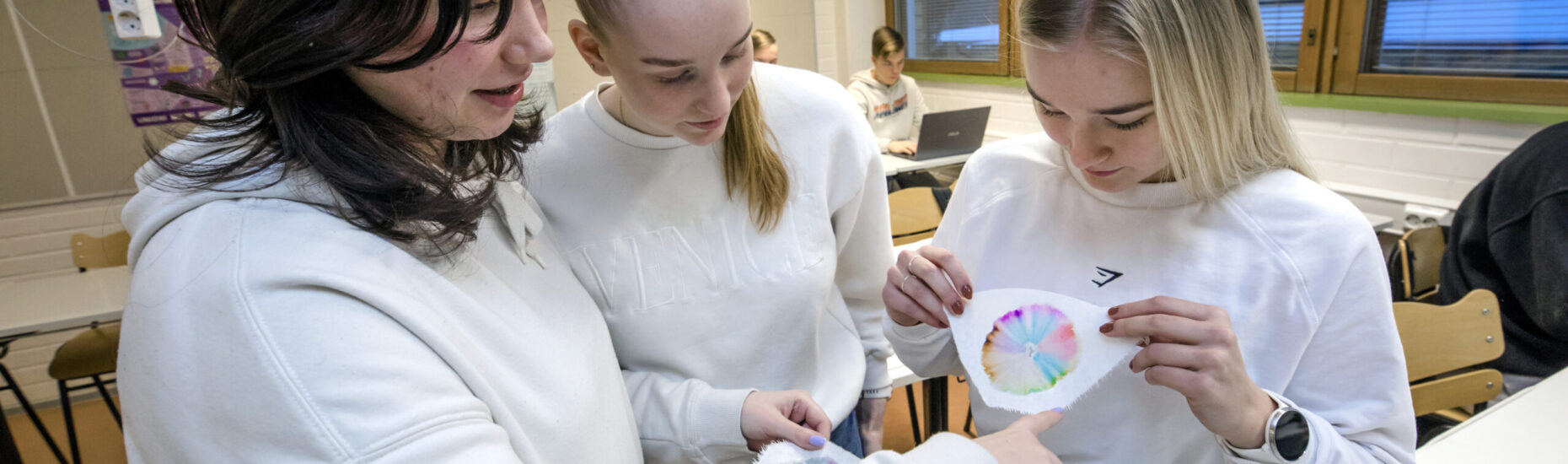 Kolme lukiolaistyttöä seisoo koululuokassa pöydän ääressä tutkimassa kuviota paperilla.