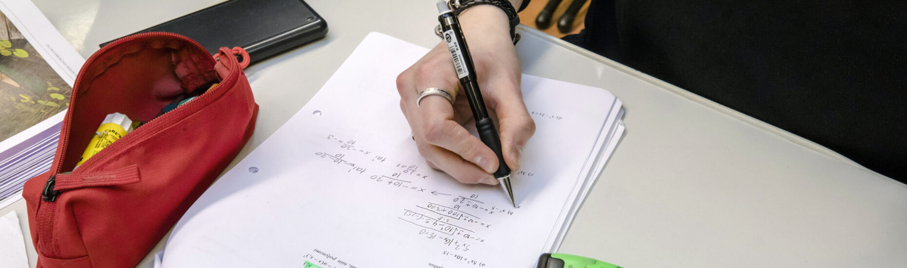 Lähikuva pulpetilta, jolla penaali, kännykkä, terotin, kaksi tussia sekä paperi, jolle käsi juuri kirjoittamassa.