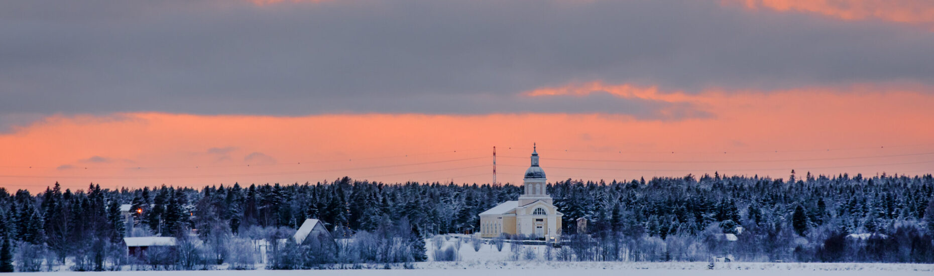 Talvinen maisema. Etualalla luminen joen jää. Vastarannalla keltainen kirkko ja pienempi vanha kivikirkko, metsää sekä oranssinkeltainen pilvien halkoma taivas.