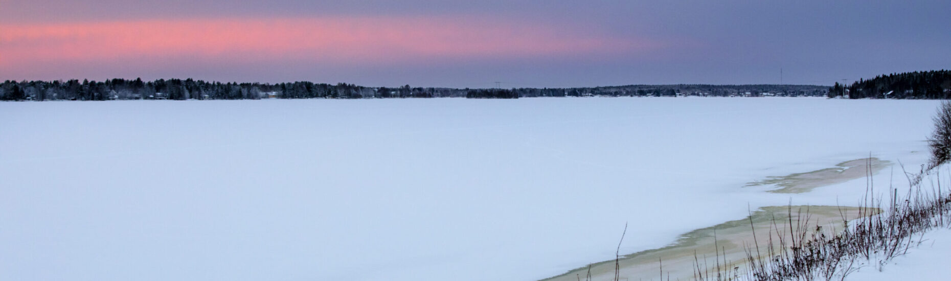 Talvinen maisema. Etualalla jäätynyt luminen joki. Vastarantaa sekä sini-punainen taivas.