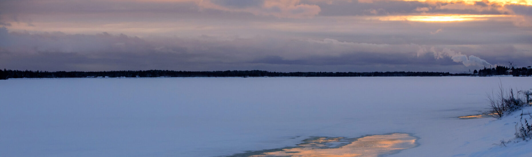 Jäinen luminen joki, tummaa vastarantaa ja keltainen taivas, jota halkovat siniharmaat pilvet.