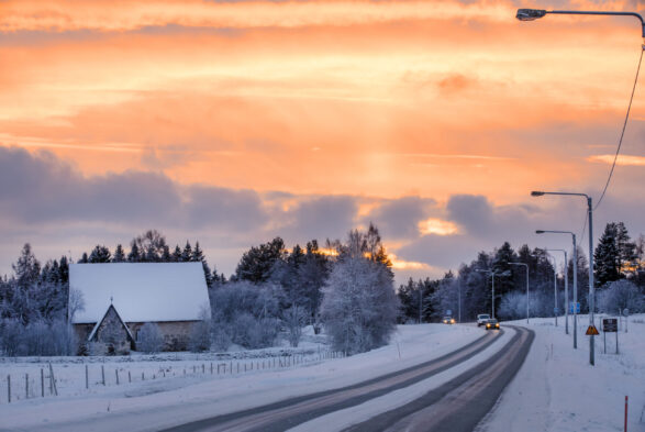 Keskiaikainen kivikirkko talvisessa maisemassa. Taivas keltainen, sinertäviä pilviä. Oikealla kirkon vieritse kulkeva tie ja autoja.